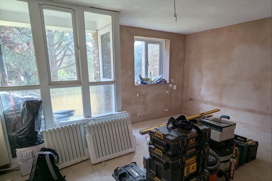 Living room at the Innes Gardens flat with fresh pink basecoat plaster on the walls, new radiators leaning against the full-height window and tool cases on the bare floor