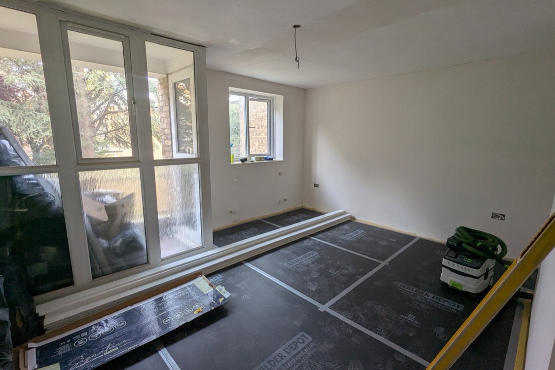 The same living room at the Innes Gardens flat after skimming, walls smooth and pale, floor covered with protection sheets and new skirting boards laid out by the window